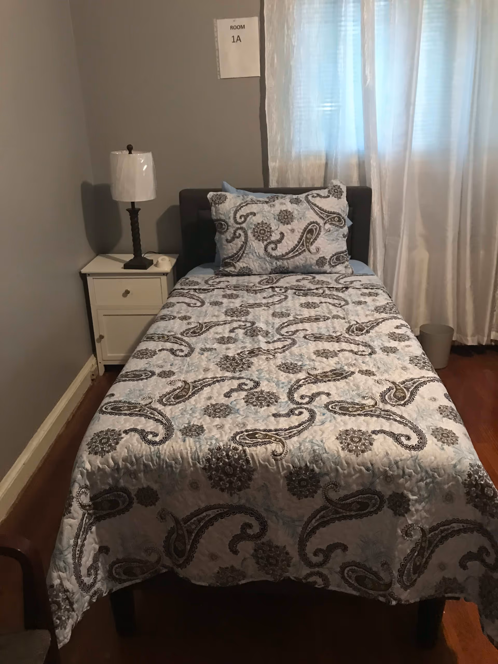 A small bedroom with a single bed covered in a patterned quilt and matching pillow. Next to the bed is a white nightstand with a lamp and a small item on it. A window with sheer white curtains is behind the bed. A sign on the wall above the bed reads 'ROOM 1A'.