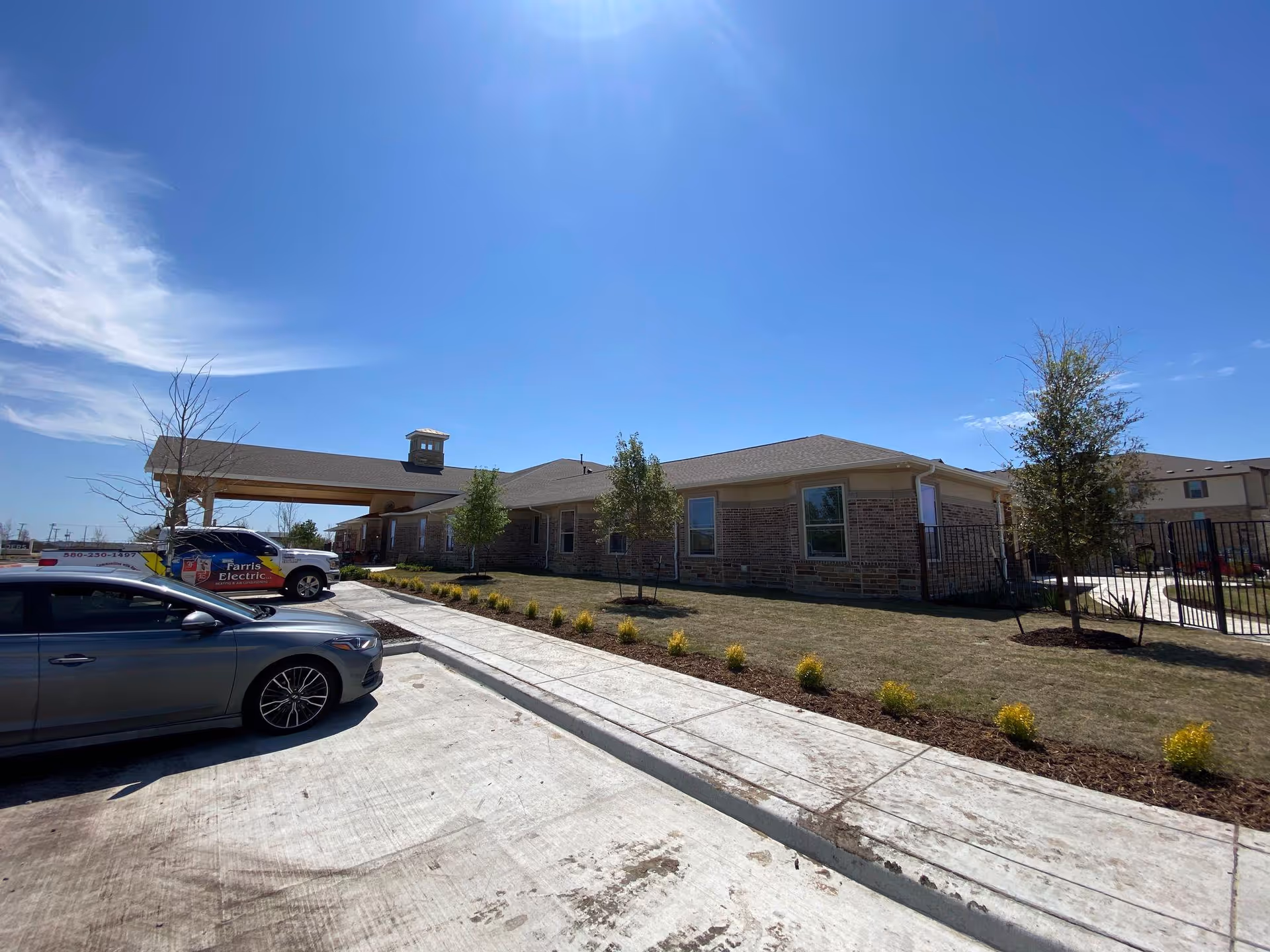 Single-story brick senior living building exterior with a covered entrance, lawn, and parked cars.