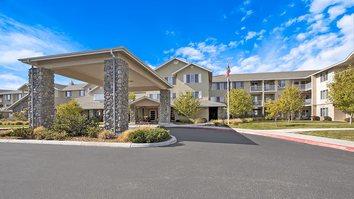 Exterior view of Holiday Sky Peaks senior living facility showing a large covered entrance supported by stone pillars, a three-story building with balconies, trees, and a flagpole with an American flag under a blue sky with scattered clouds.