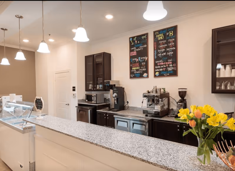Interior view of a coffee bar area with a granite countertop, dark wood cabinets, coffee machines, a microwave, and two colorful menu boards on the wall. There are pendant lights hanging from the ceiling and a vase with yellow and orange flowers on the counter.