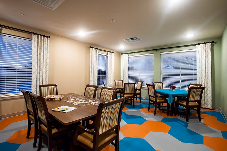 A brightly lit room with multiple wooden tables and chairs arranged for group seating. The floor features a colorful geometric pattern with orange, blue, and gray hexagons. The walls are painted beige and green, and there are large windows with white blinds and patterned curtains. One table has a blue tablecloth and a small flower arrangement, while another table has a partially completed puzzle and a book on it.
