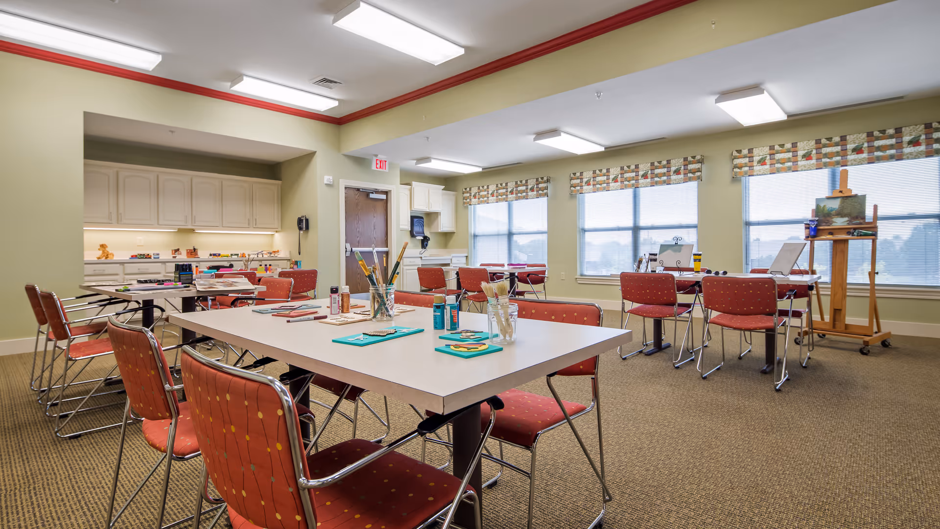 A bright activity room with several tables and red cushioned chairs arranged around them. The tables have art supplies including paintbrushes, paints, and paper. Large windows with patterned valances let in natural light. There is an easel with a painting near the windows and cabinets with additional supplies along one wall.