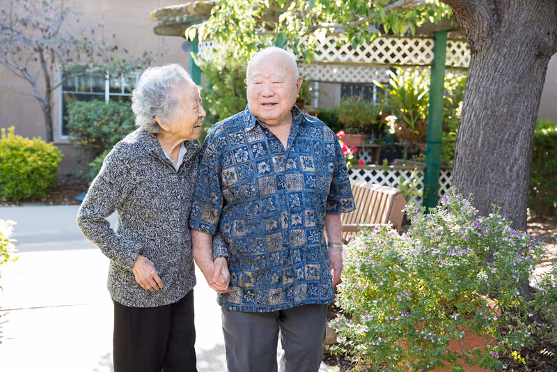 An elderly couple holding hands and walking outside in a garden area with greenery, flowers, and a tree nearby. The woman is wearing a gray sweater and black pants, and the man is wearing a blue patterned shirt and gray pants. They appear happy and engaged with each other.