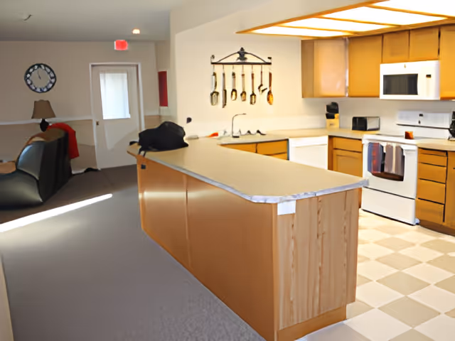 Open-plan kitchen with a long island, stove, microwave and cabinets opening into a living area with chairs and a wall clock.