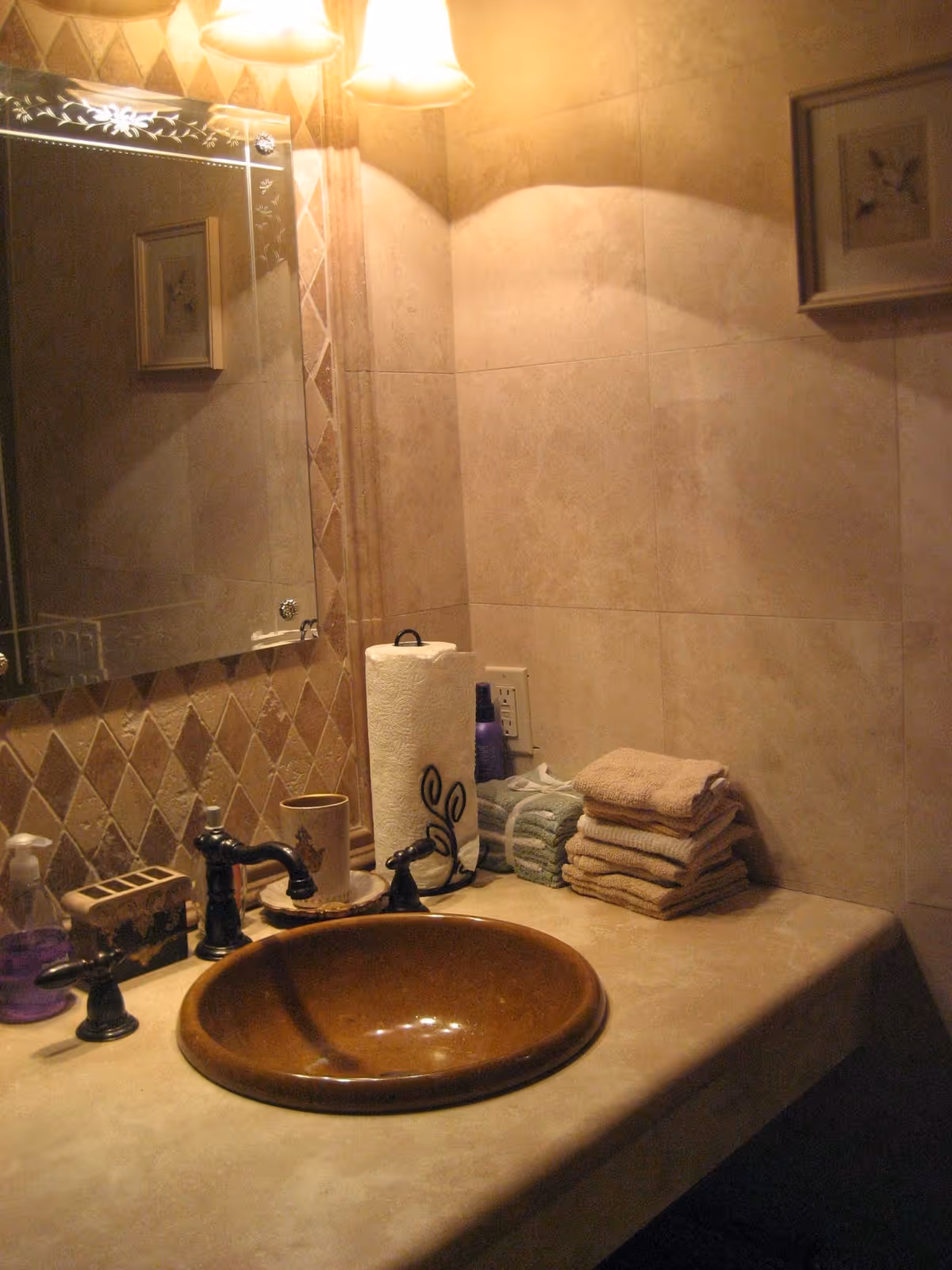 Bathroom countertop with a brown vessel sink, faucets, mirror, stacked towels and a paper towel roll under wall sconces.