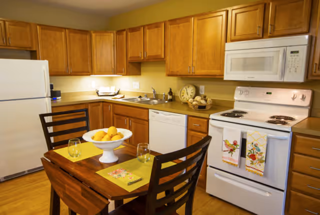 A cozy kitchen with wooden cabinets, a white refrigerator, a white microwave above a white stove, and a dishwasher. A small wooden dining table with two chairs is set with yellow placemats, two glasses, a floral napkin, and a white bowl filled with lemons. The countertop has a decorative clock, a basket with rolls, and a soap dispenser near the sink.