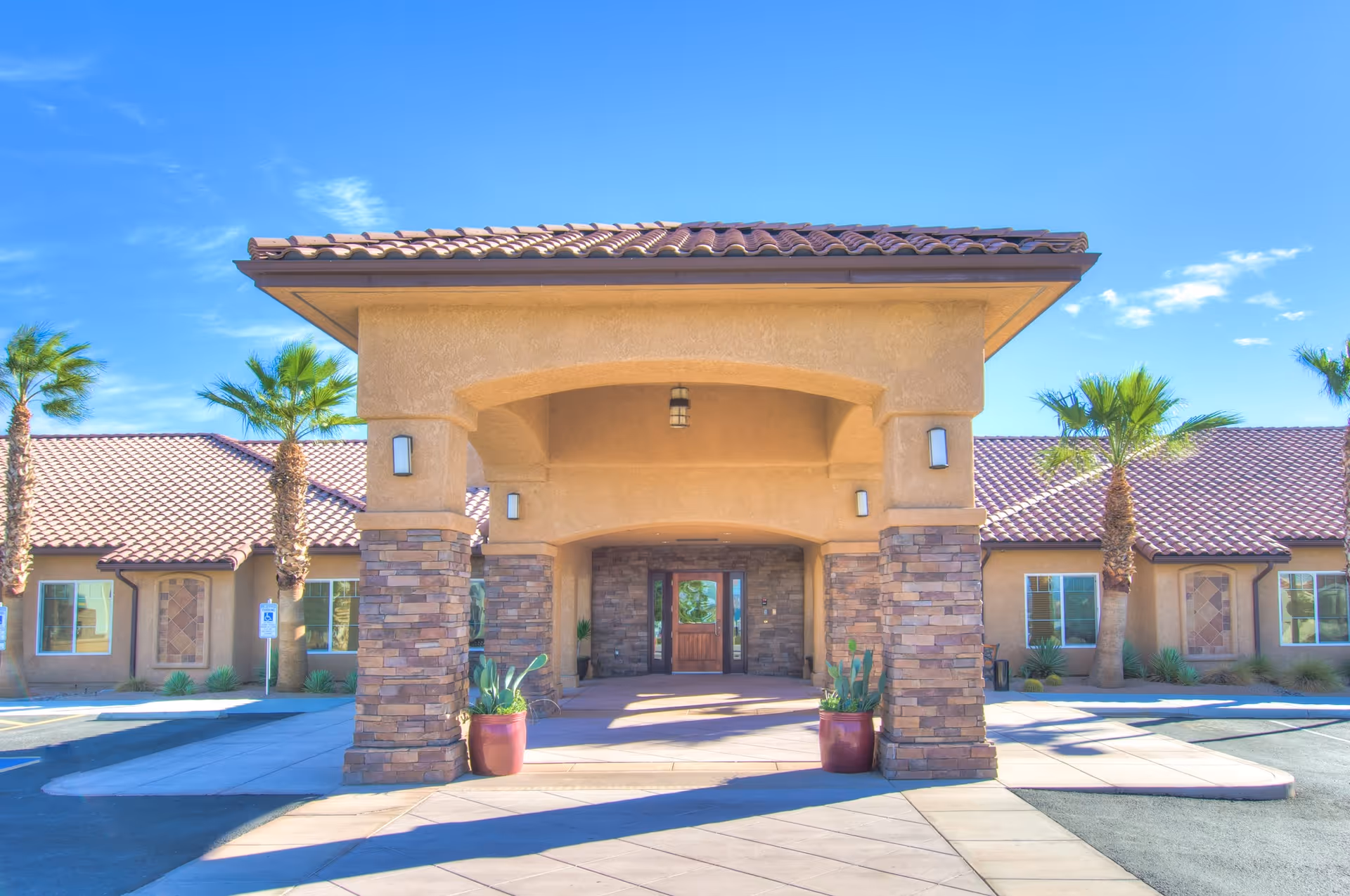 Front exterior view of Lake View Terrace Memory Care Residence featuring a covered entrance with stone pillars, potted plants, palm trees, and a clear blue sky.