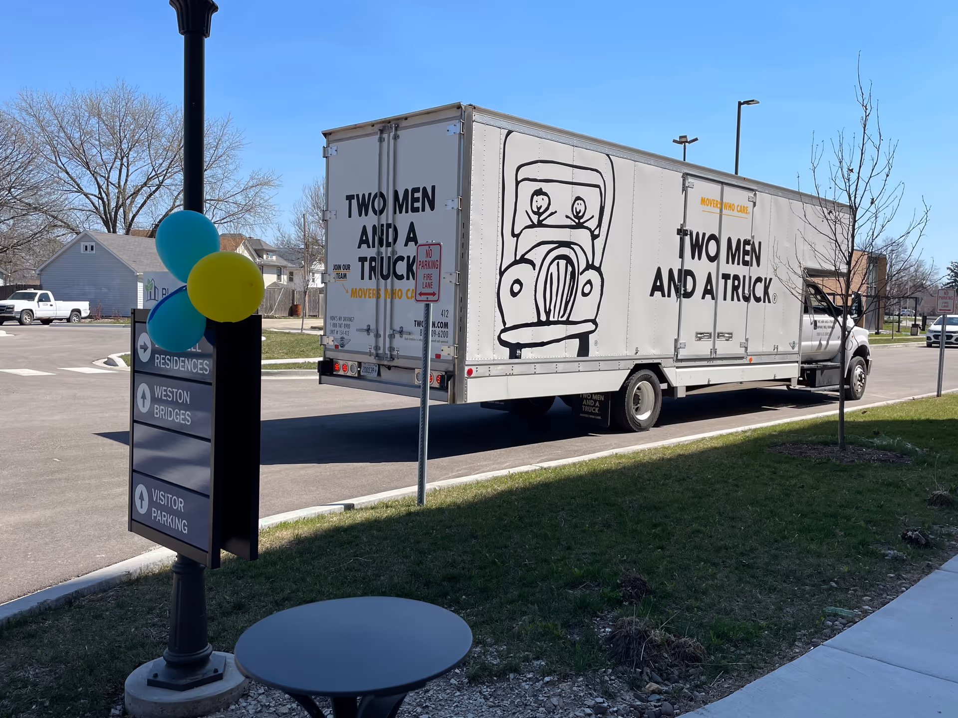 A large 'Two Men and a Truck' moving truck parked on the street outside the Bardwell Residences with a signpost and balloons in the foreground.