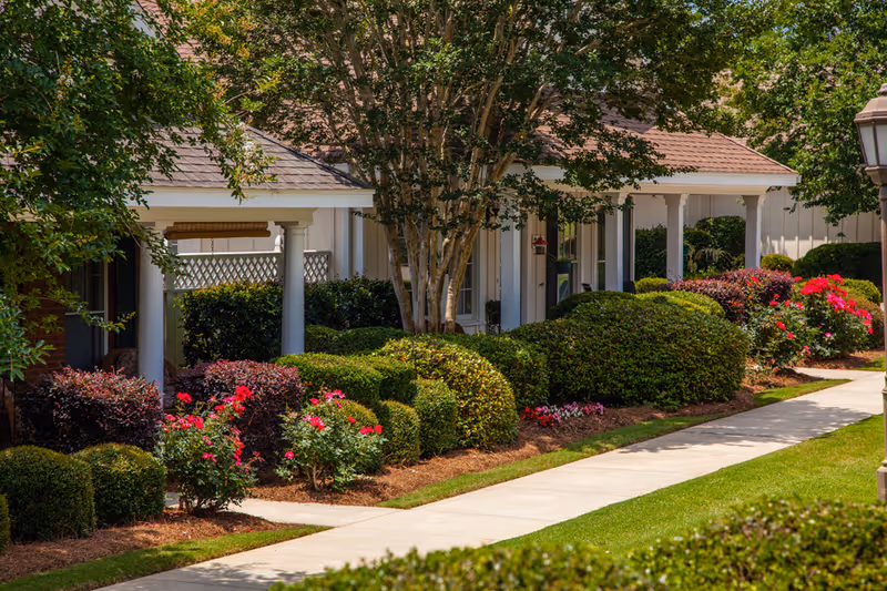 Walkway flanked by manicured hedges and flowering shrubs leading to the porch of a senior living building.