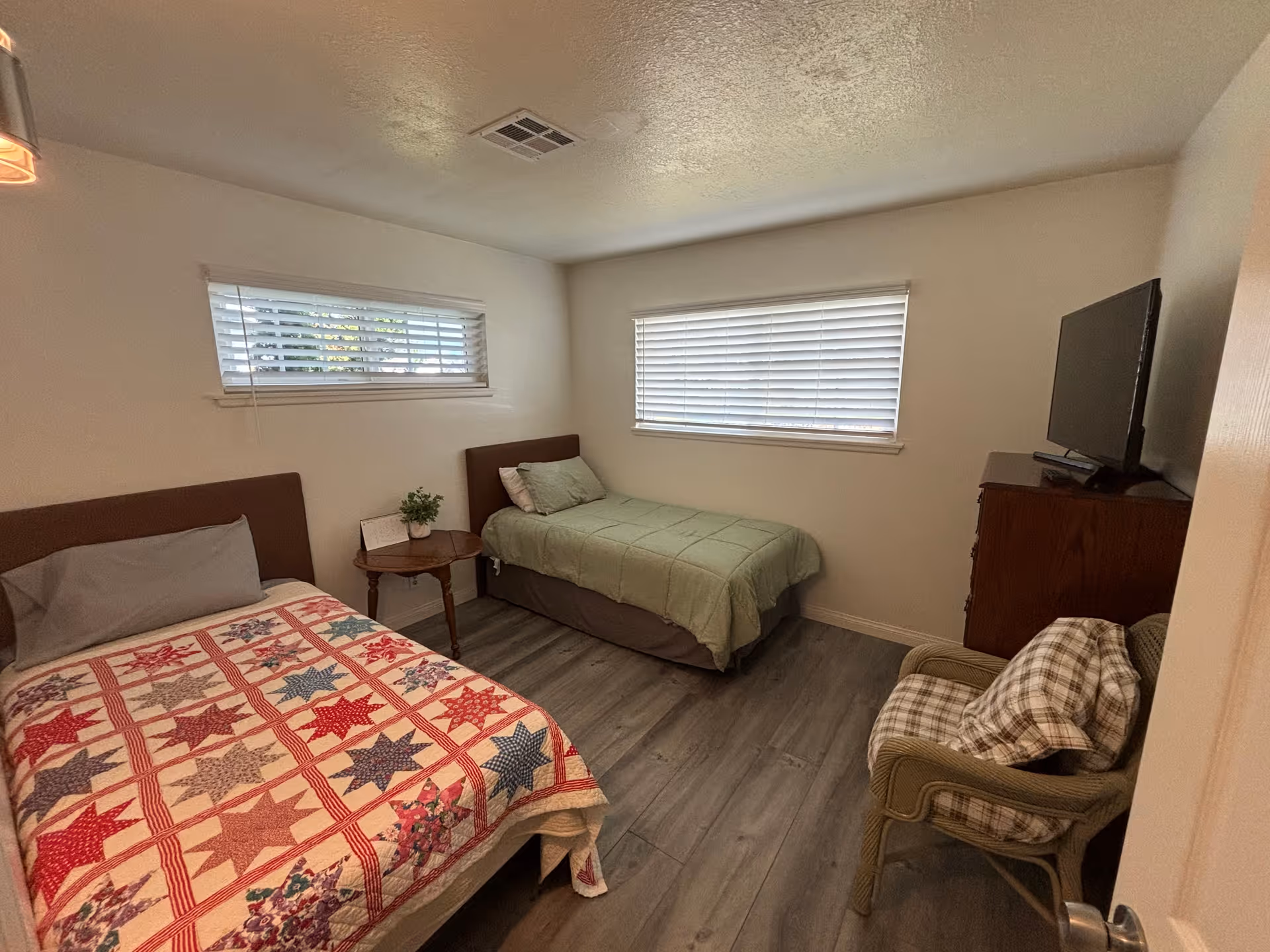 Bedroom with two single beds, a small bedside table, dresser with a TV, and a chair under two horizontal windows.