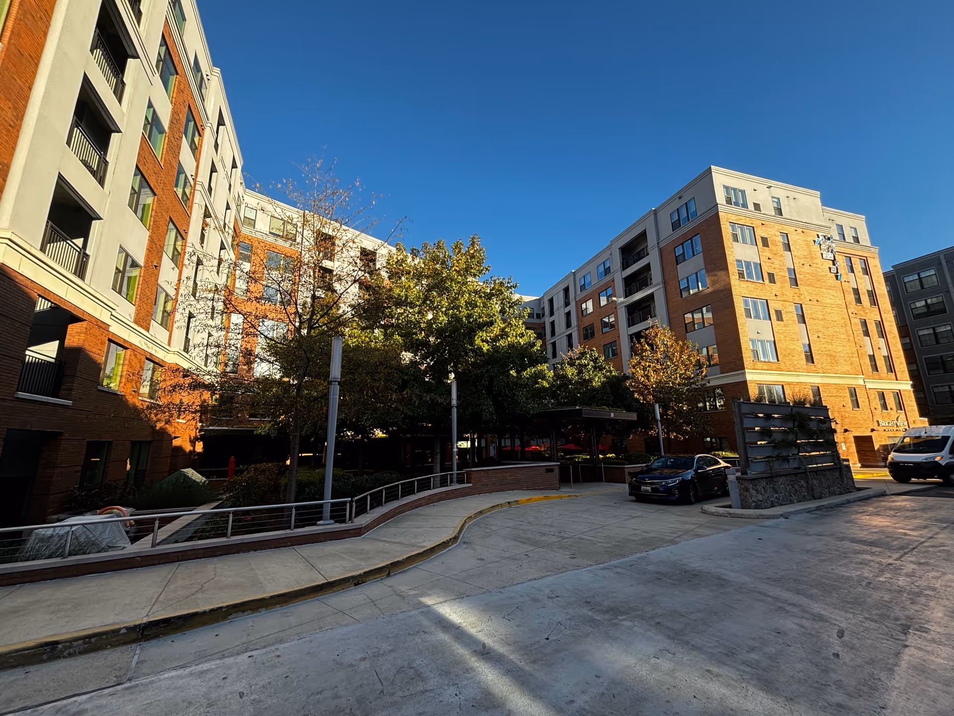 Exterior view of a multi-story senior living facility building with brick and light-colored walls, surrounded by trees and a driveway with parked cars under a clear blue sky.