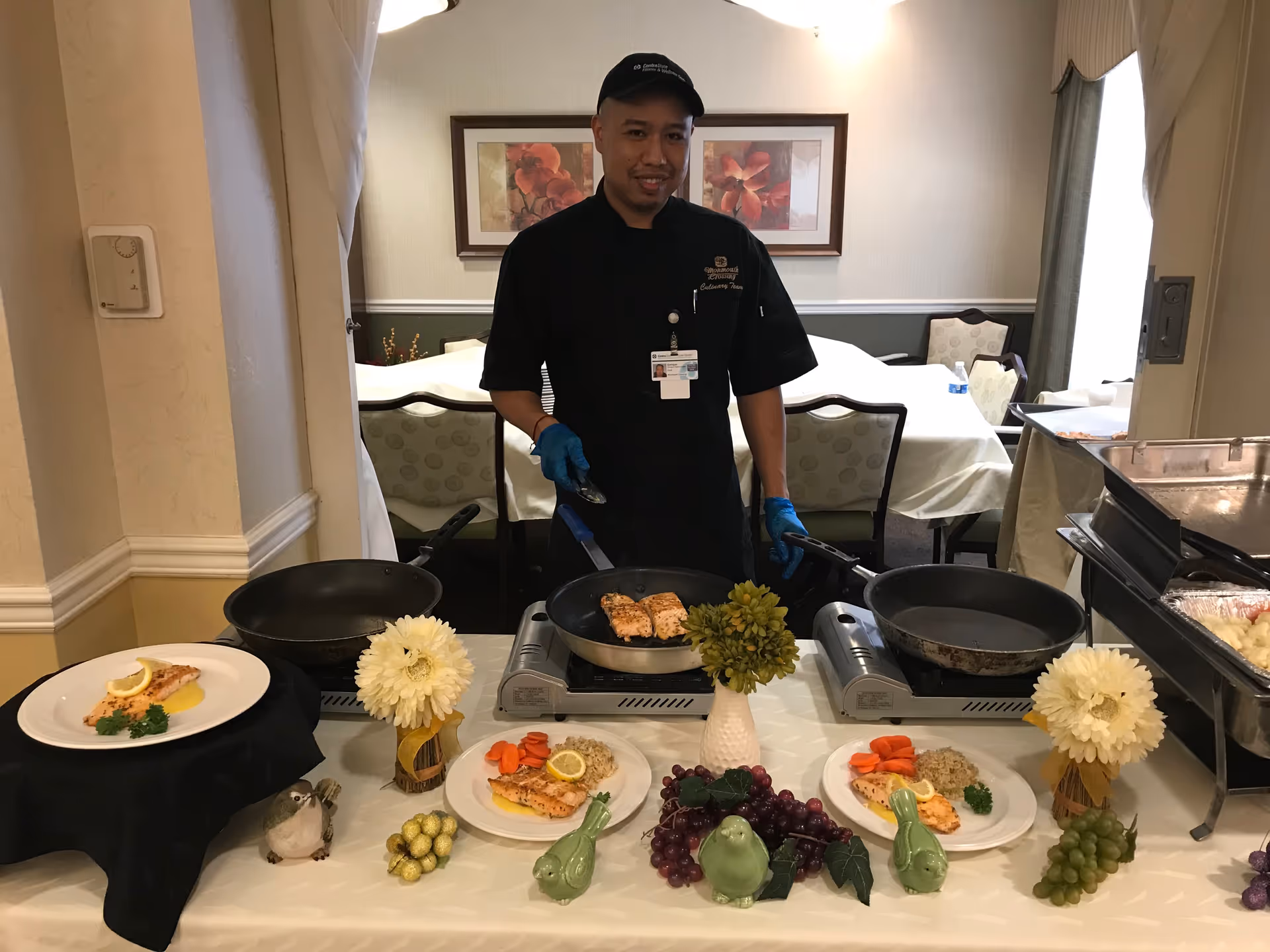 A chef wearing a black uniform and blue gloves stands behind a table with three portable stoves, cooking salmon fillets in a pan. The table is decorated with flowers, ceramic birds, and artificial grapes. Plates with cooked salmon, carrots, and rice are arranged on the table. The background shows a dining area with chairs and tables covered with white tablecloths.