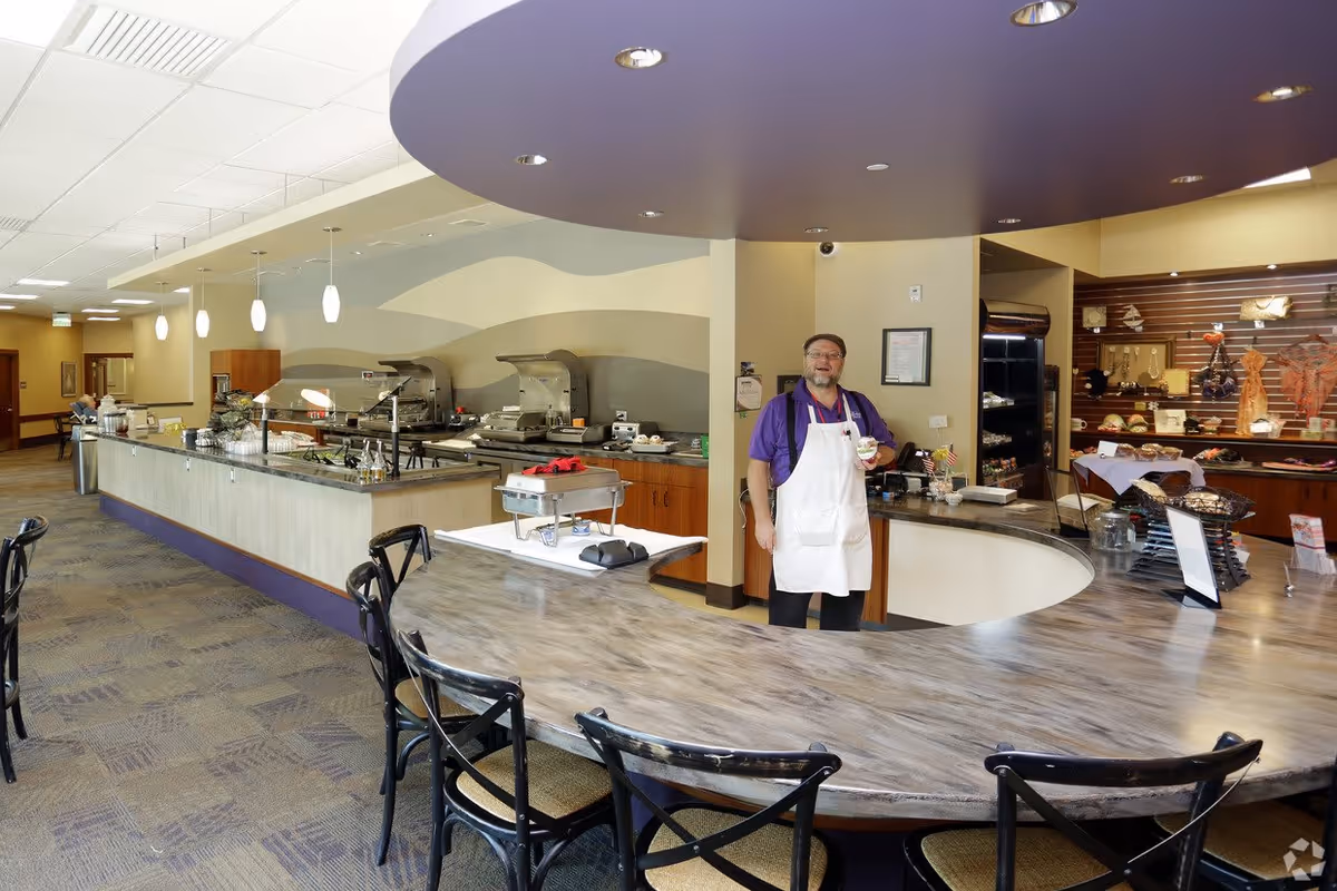 A man wearing a purple shirt and white apron stands behind a curved counter in a dining area with a buffet setup. The room has a modern design with pendant lights hanging over the buffet, various food items displayed, and several black chairs arranged around the counter.