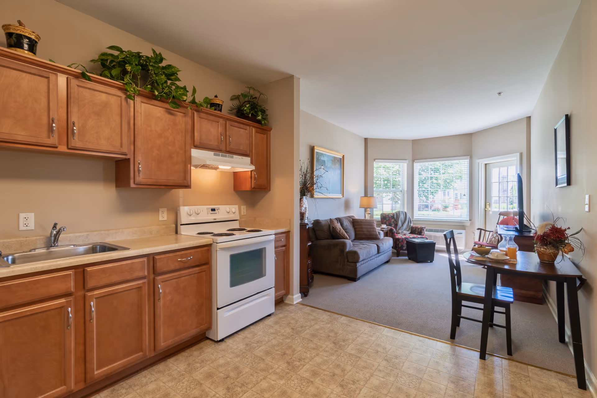 Open-plan interior showing a kitchen with wooden cabinets and a white stove opening into a living area with a sofa, dining table, and bay windows.