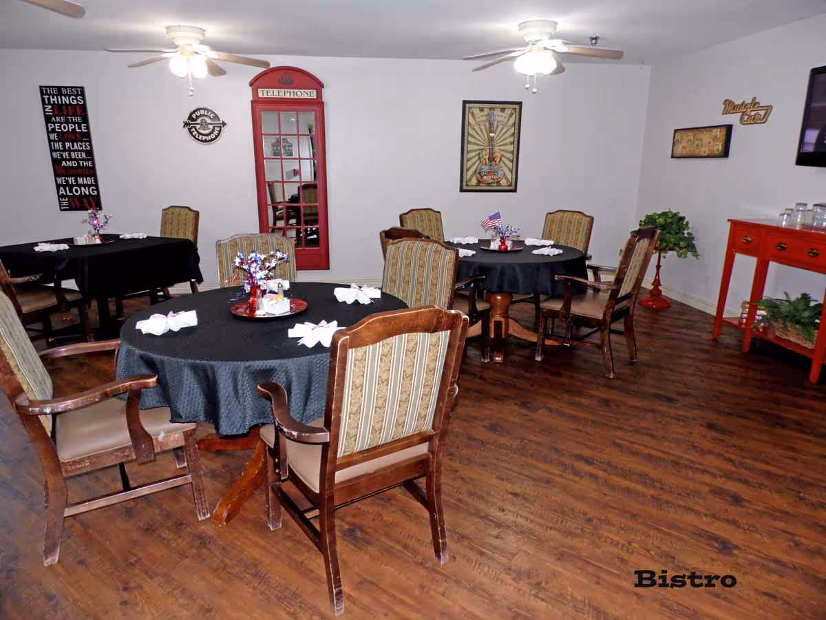 Interior view of a dining area with round tables covered in black tablecloths, each surrounded by wooden chairs with patterned cushions. The room has wooden flooring, ceiling fans with lights, and various wall decorations including a red telephone booth-style cabinet, framed pictures, and a sign that says 'Bistro'.