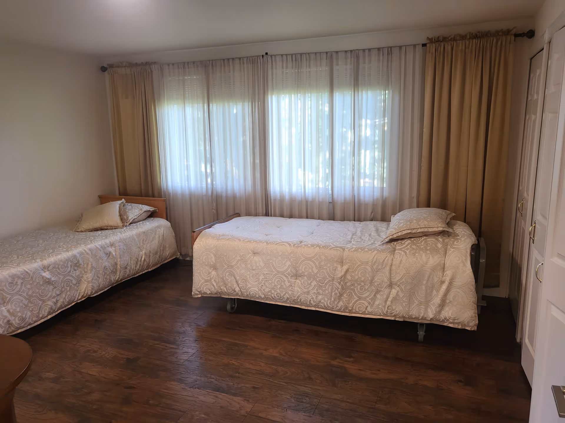 A bedroom with two single beds covered in beige patterned bedspreads, each with a matching pillow. The room has wooden flooring, a large window with sheer white curtains and beige drapes, and a white closet with gold handles on the right side.