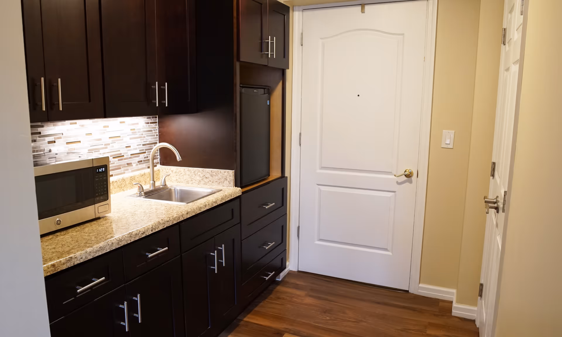 Small kitchenette with dark cabinets, granite countertop, stainless sink and microwave next to a white door.