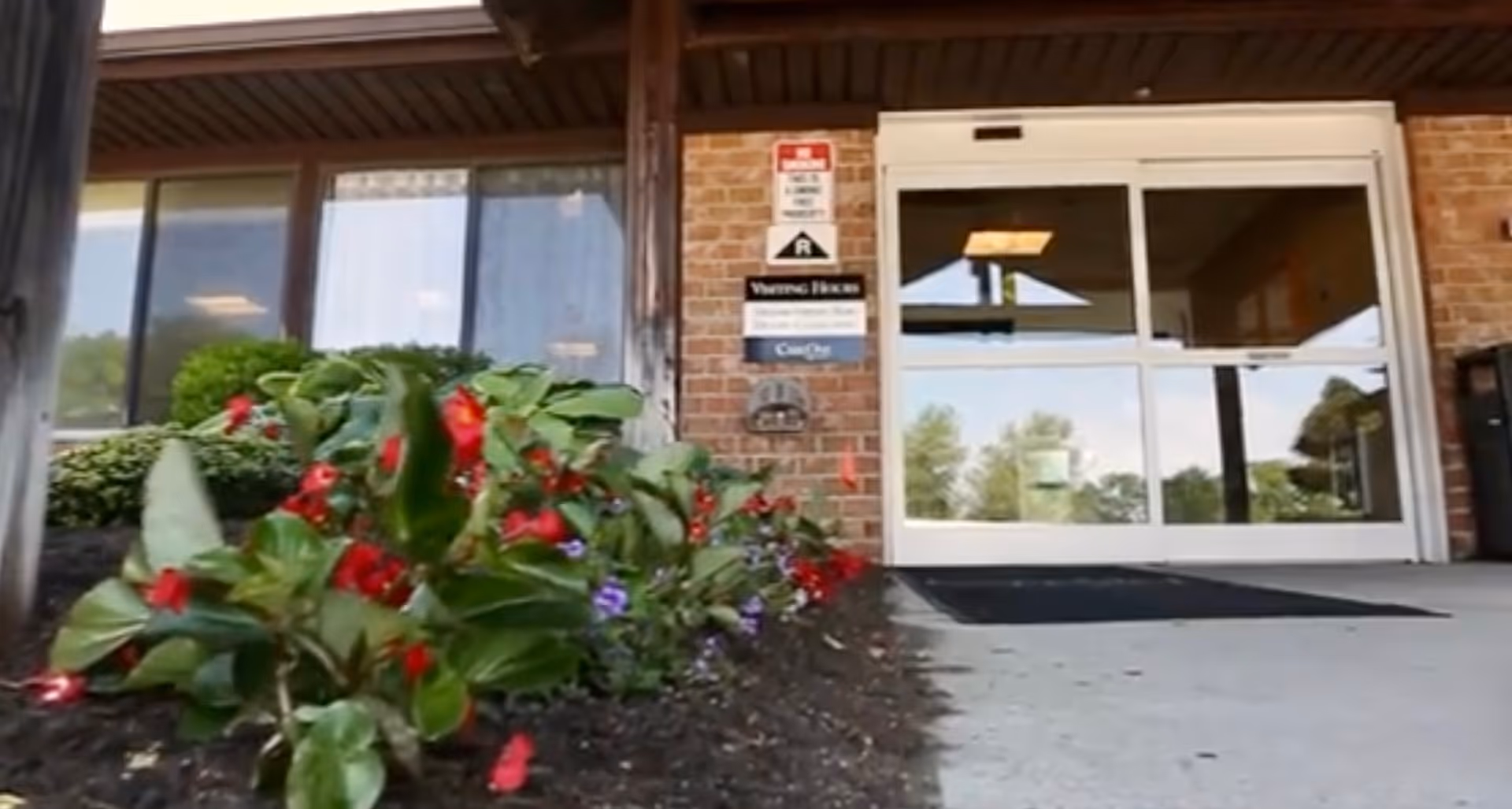 Brick building entrance with glass doors and a flowering garden in front.