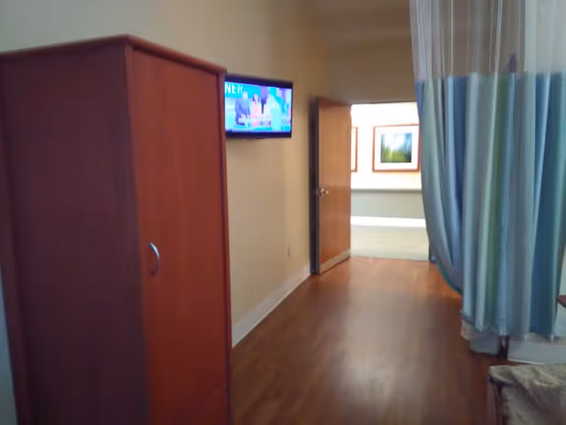 Interior view of a rehab room showing a wooden cabinet, wall-mounted TV, privacy curtain, and an open door to a hallway.