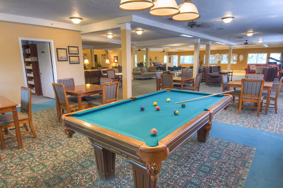Indoor common room with a pool table in the foreground and tables, chairs, and lounge seating in the background.