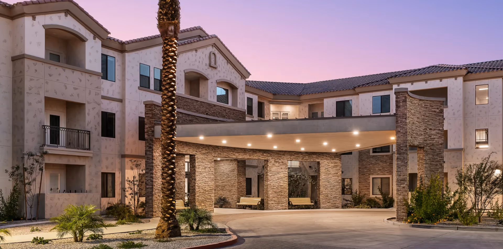 Exterior view of Cadence Chandler senior living facility at dusk, showing a three-story building with beige walls, stone accents, a covered entrance with lights, palm trees, and landscaped greenery.