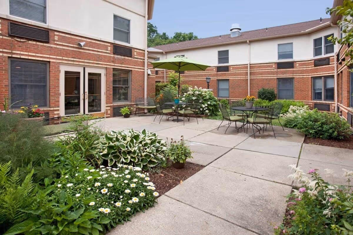 Outdoor courtyard area at The Pines of Mount Lebanon featuring paved walkways, green plants, flowers, and patio tables with chairs and umbrellas surrounded by a two-story brick and beige building.