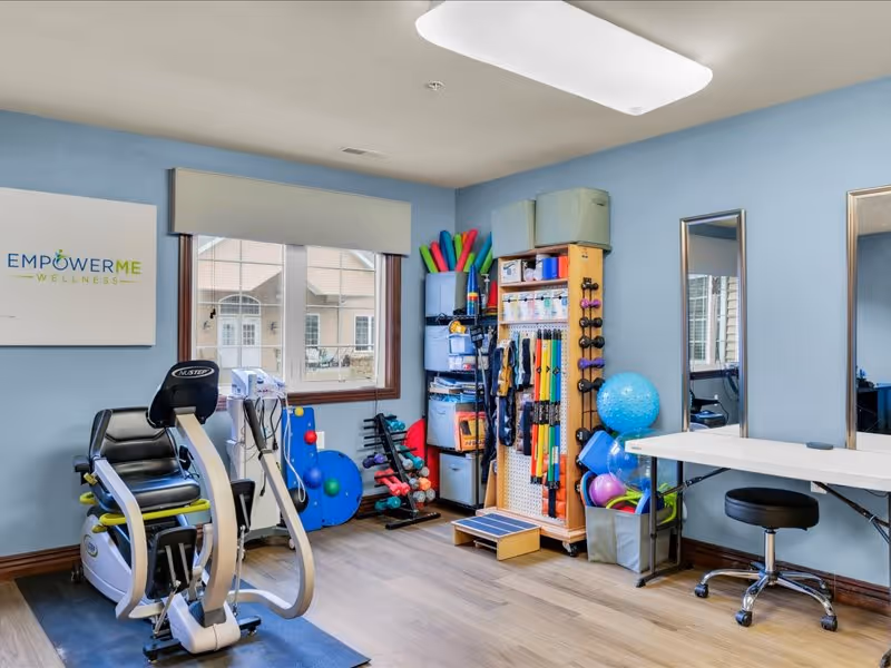 A wellness room with exercise equipment including a recumbent bike, various fitness accessories such as resistance bands, weights, exercise balls, and foam rollers organized on shelves and racks. The room has light blue walls, a window with a view outside, two mirrors on the wall, and a white table with a black swivel chair.