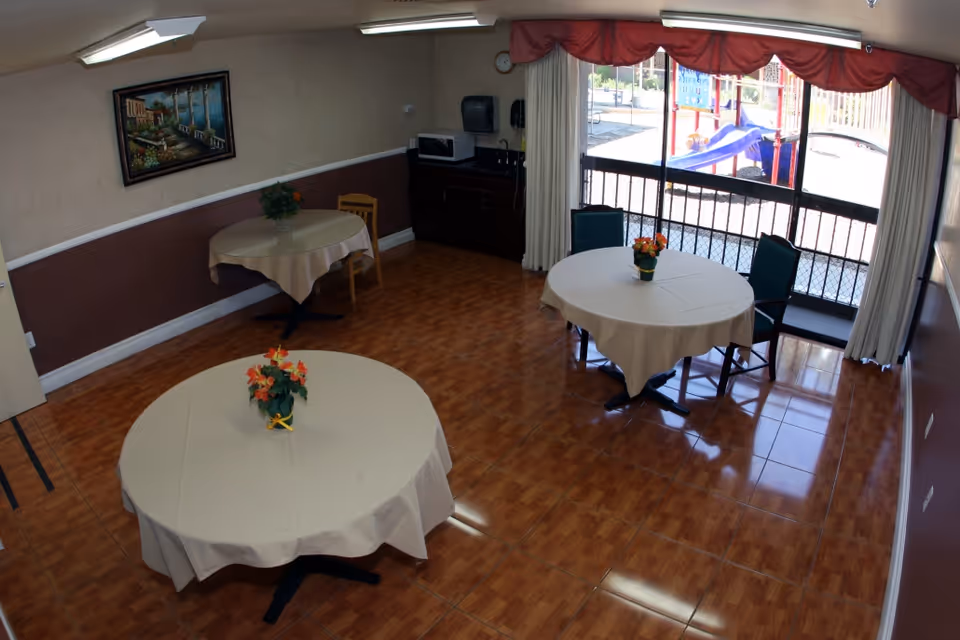 Small dining/activity room with round tables covered in white tablecloths, chairs, floral centerpieces, and large windows overlooking an outdoor playground.