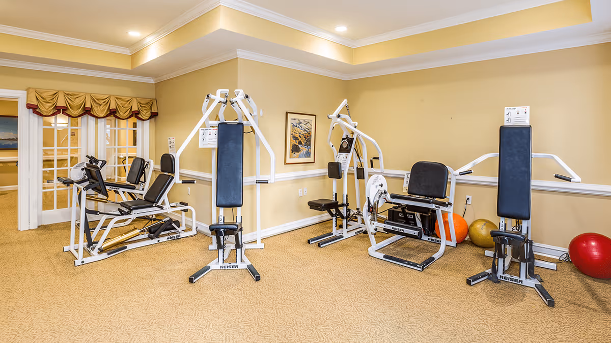A small fitness room with beige walls and carpeted floor containing several pieces of white exercise equipment including weight machines and a leg press. There are colorful exercise balls in the corner and a framed picture on the wall. The room has a tray ceiling with recessed lighting and a window with a valance.