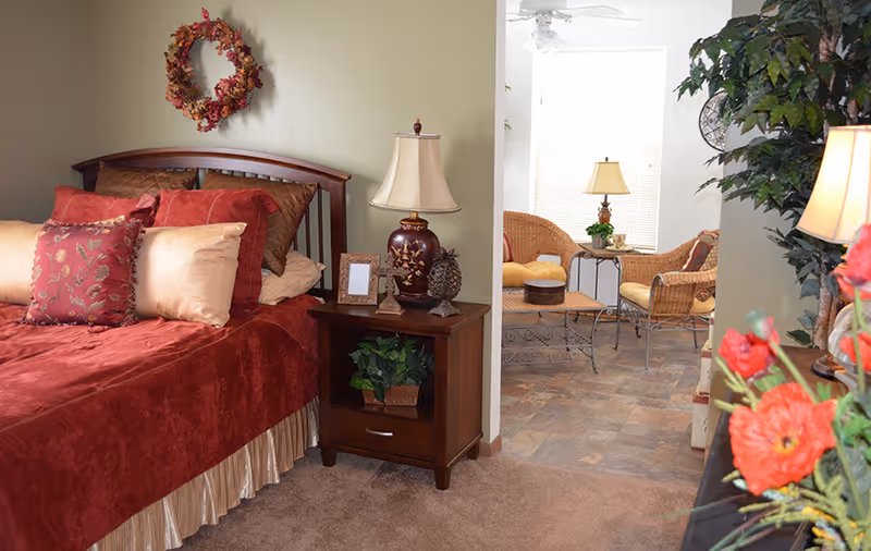 Cozy bedroom with a red bedspread, wooden nightstand and lamp, opening into a sunlit seating area with wicker chairs.