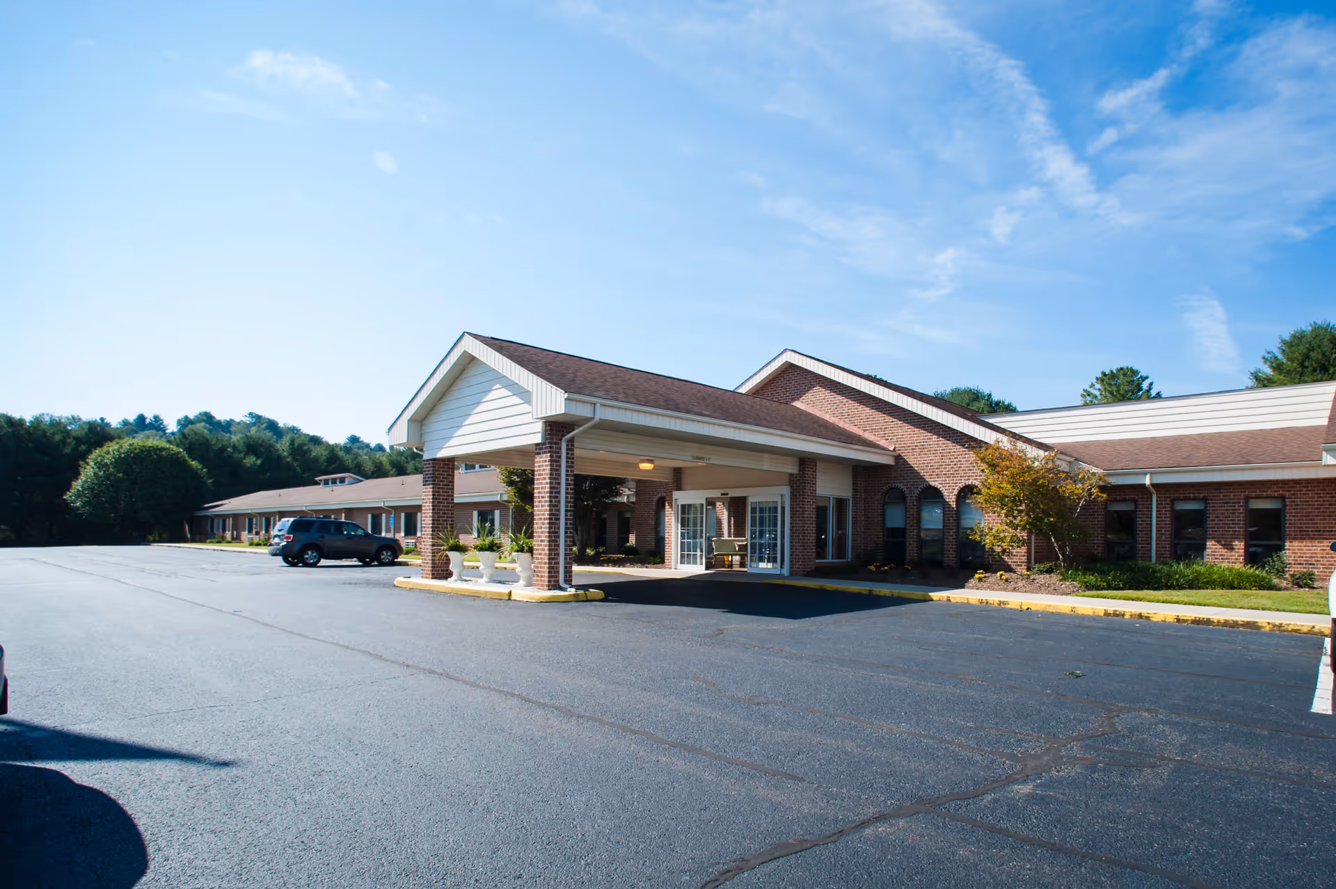 Exterior view of a single-story brick building with a covered entrance supported by brick columns. There are a few cars parked in the paved parking lot in front of the building, and trees and greenery are visible in the background under a clear blue sky.