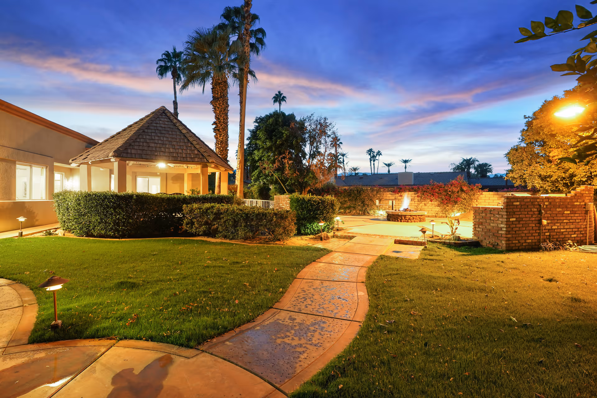 Outdoor area of a senior living facility at dusk featuring a paved walkway, well-maintained green lawn, palm trees, a small gazebo with a shingled roof, and a brick fire pit surrounded by a low brick wall. The sky is a mix of blue and purple hues with some clouds, and the area is softly illuminated by outdoor lighting.