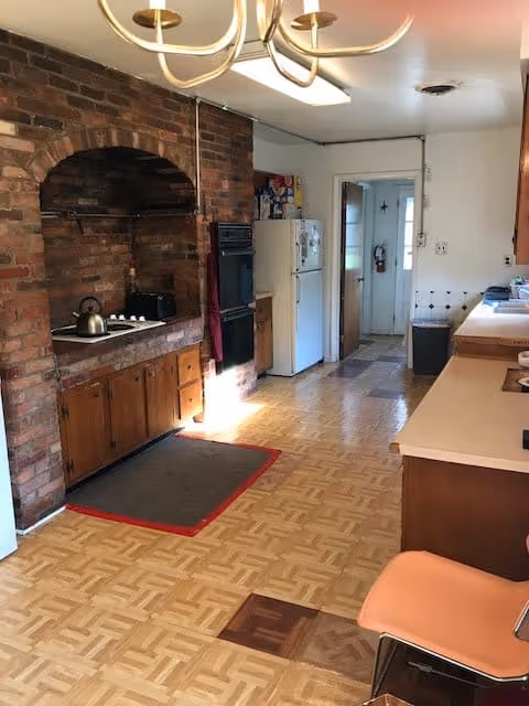 A long kitchen with a brick cooking alcove and stovetop on the left, a refrigerator and oven at the far end, and countertops and seating along the right.