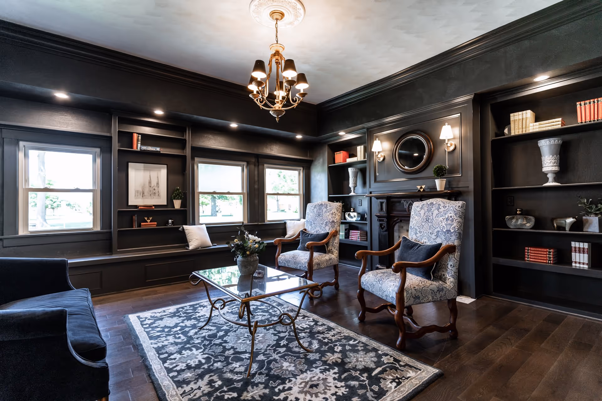 A cozy living room with dark walls and wood flooring featuring two patterned armchairs with dark cushions, a glass coffee table with a decorative plant, a dark blue sofa, built-in shelves with books and decorative items, three windows letting in natural light, and a chandelier hanging from the ceiling.