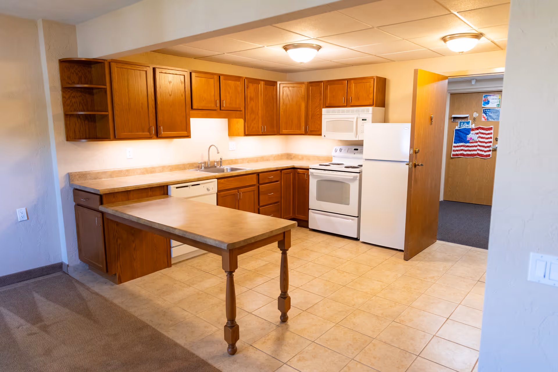 Small kitchen with wooden cabinets, white appliances, a central peninsula table, and tiled floor in a senior living apartment.