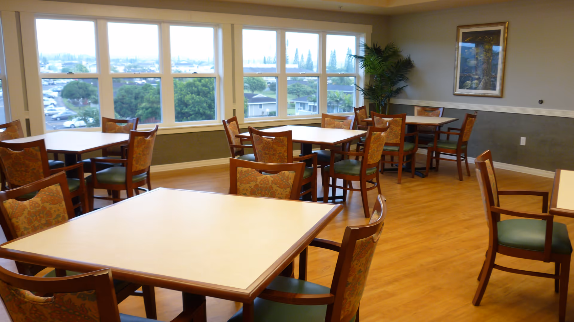 A bright dining room with several square tables and wooden chairs with patterned cushions. Large windows provide a view of trees, houses, and a parking lot outside. The room has wooden flooring, a framed picture on the wall, and a tall green plant in the corner.