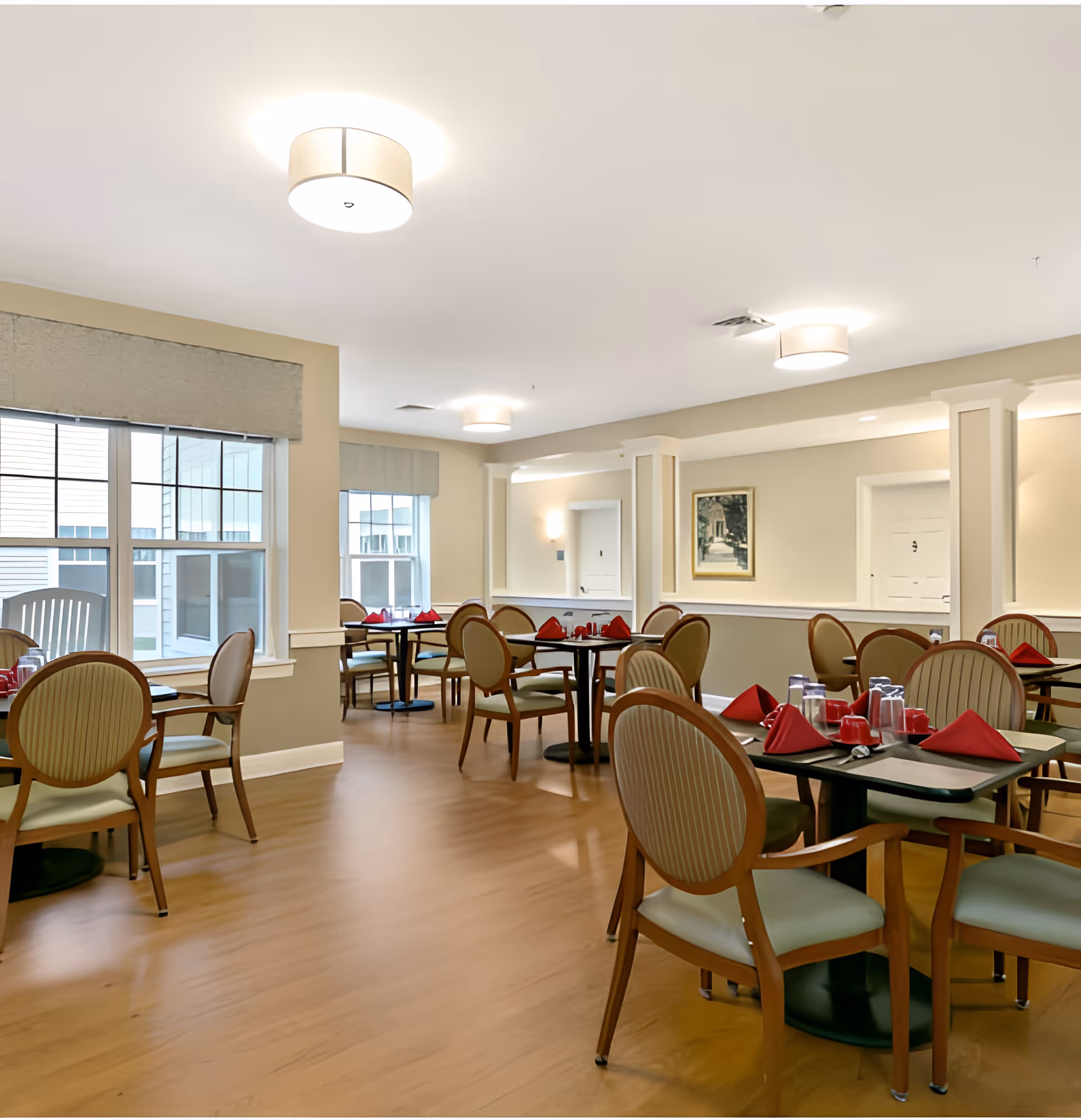A bright and spacious dining room in an assisted living facility with multiple round tables set with red napkins, glasses, and utensils. The room features wooden chairs with cushioned seats and backs, large windows with beige valances allowing natural light, light-colored walls, and modern ceiling lights.