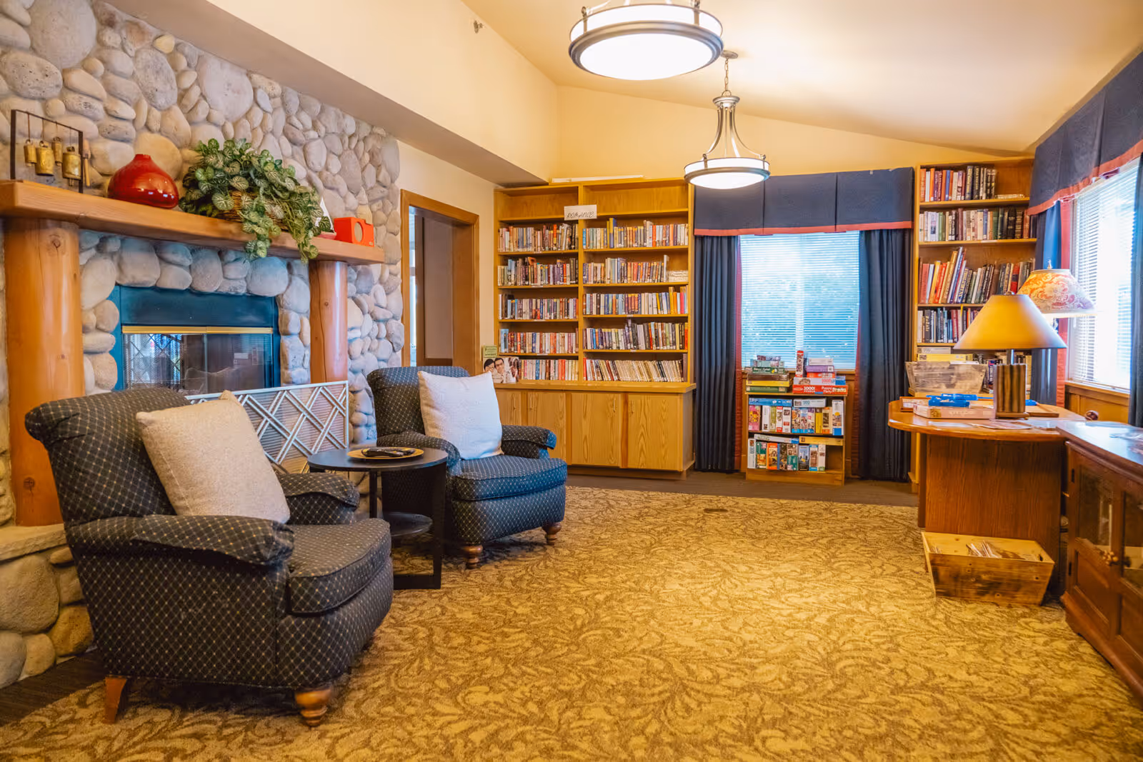 Cozy living room area with two patterned armchairs and a small round table in front of a stone fireplace. The room has carpeted flooring, wooden bookshelves filled with books, a window with blue curtains, and a wooden desk with a lamp and board games stacked on a shelf nearby.