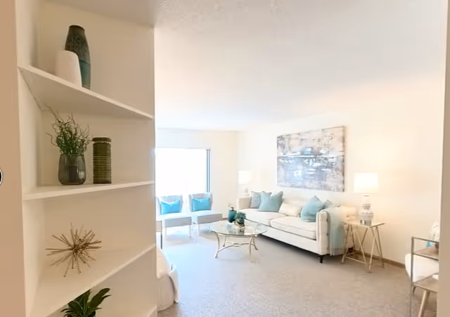 Bright, softly furnished living room with a white sofa, glass coffee table, accent chairs, and decorative shelving.