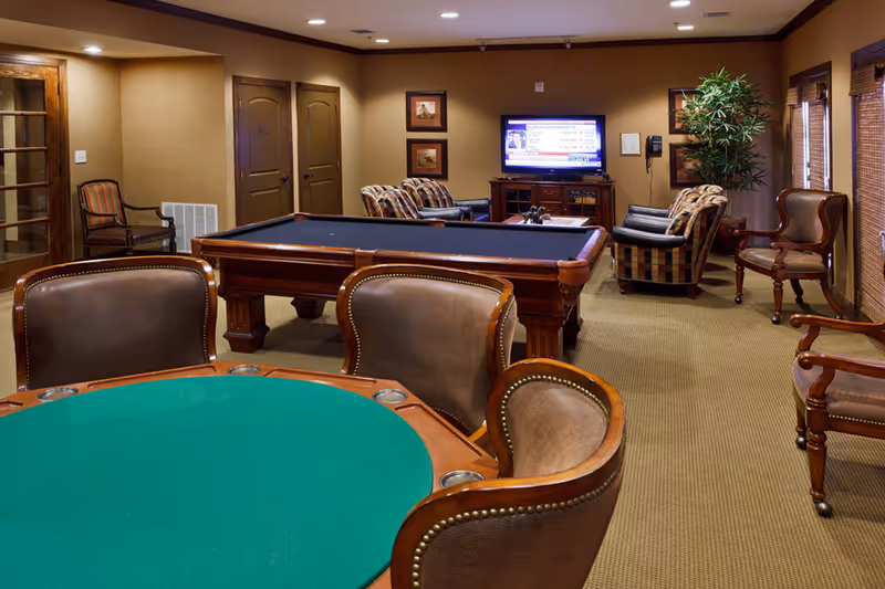 A cozy game room with a green felt poker table surrounded by leather chairs in the foreground, a pool table in the middle, and a seating area with armchairs and a TV mounted on the wall in the background. The room has warm lighting, beige walls, and carpeted flooring.