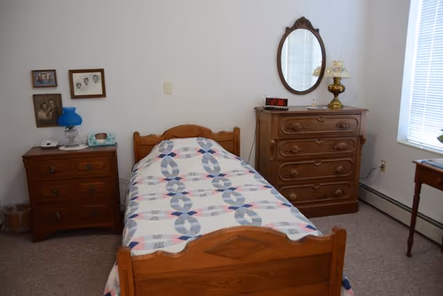 A small bedroom with a single wooden bed covered with a patterned quilt. To the left of the bed is a wooden nightstand with a blue lamp, a telephone, and framed photos on the wall above it. To the right of the bed is a wooden dresser with an oval mirror, a vintage lamp, and a digital clock showing 2:36. A window with blinds is on the right wall, letting in natural light.