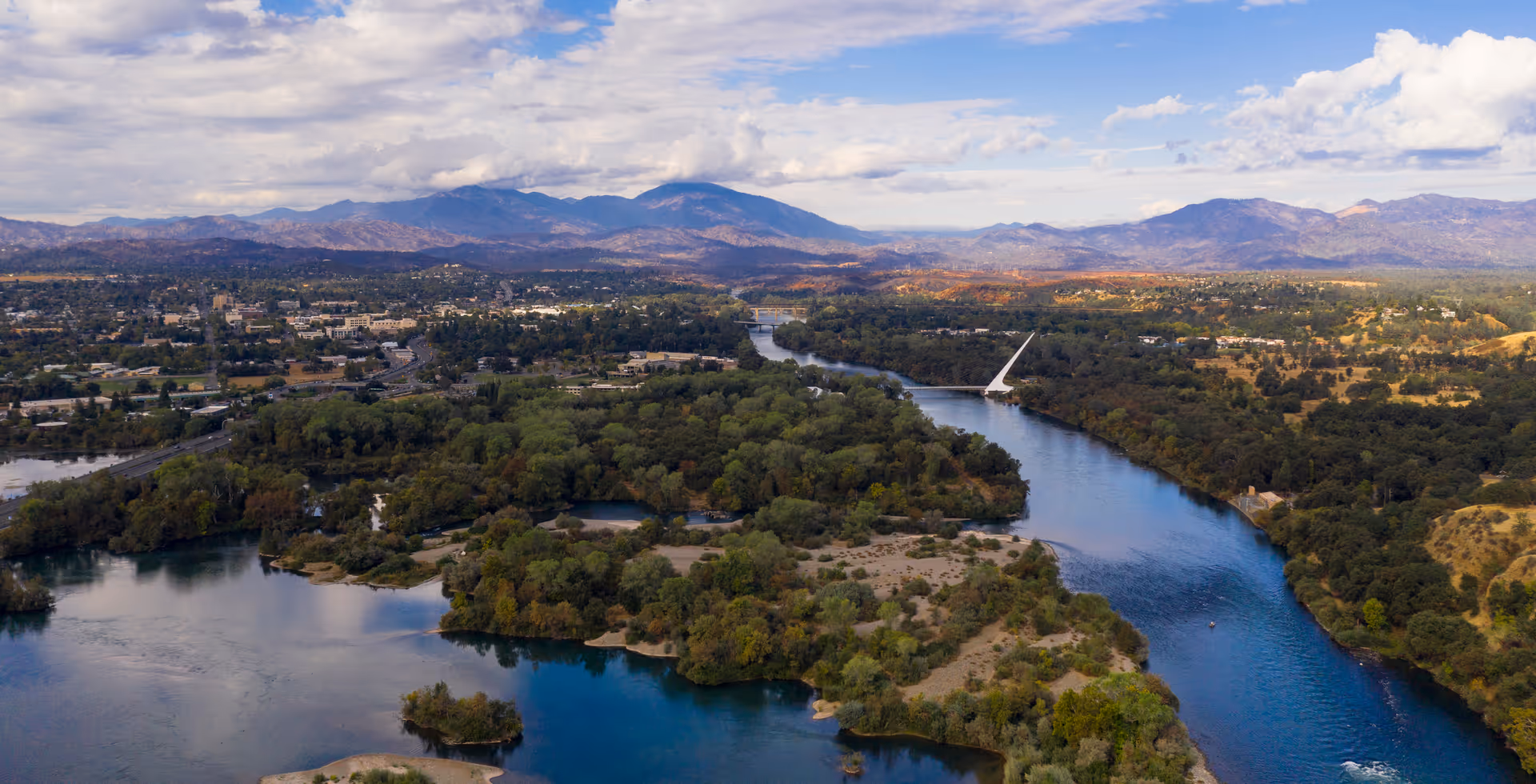 Aerial view of a river winding through forested islands with a city and mountains in the background.