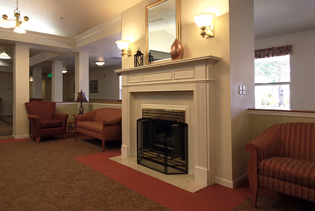 A cozy living room area in a senior living facility featuring a white fireplace with a mirror and decorative items on the mantel. There are upholstered chairs and sofas arranged around the room, with soft lighting from wall sconces and ceiling fixtures. A window with a floral valance lets in natural light.