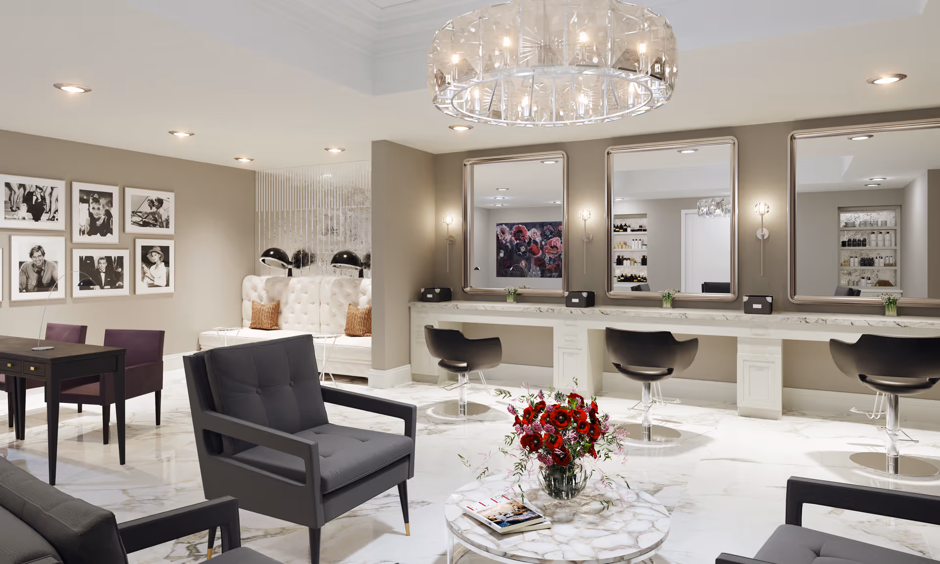 A modern and elegant salon area with three styling stations featuring large mirrors and black swivel chairs. The room has a marble floor, a round marble table with a vase of red flowers, and comfortable gray armchairs. On the left side, there is a small table with two purple chairs and a wall decorated with black and white framed photographs. The lighting includes recessed ceiling lights and a large decorative chandelier.
