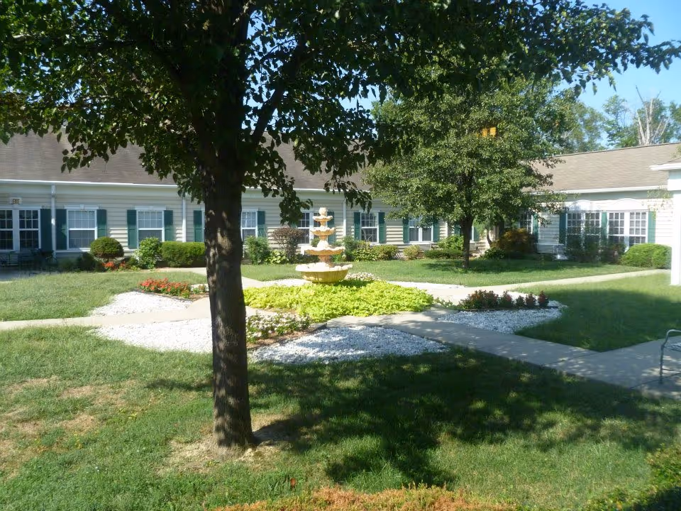 Courtyard of a senior living facility with a central multi-tiered fountain, trees, walkways, and surrounding single-story buildings.