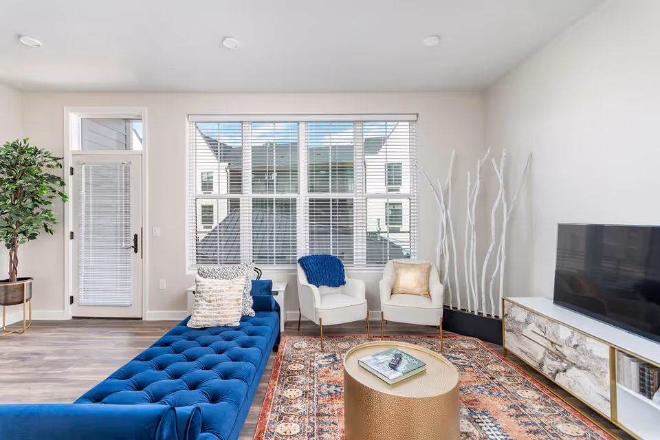 Bright living room with a blue tufted sofa, two white armchairs, large window, TV and decorative rug.