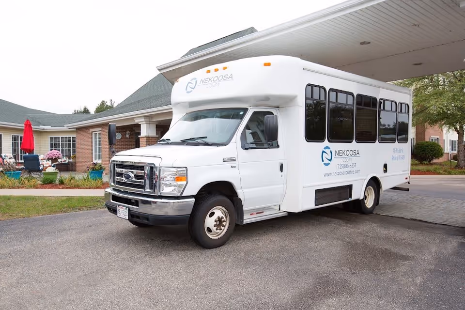 A white shuttle bus parked outside a senior living facility named Nekoosa Court. The bus has the Nekoosa Court logo and contact information on its side. The building behind the bus has a brick exterior with a covered entrance and some outdoor seating with a red umbrella.