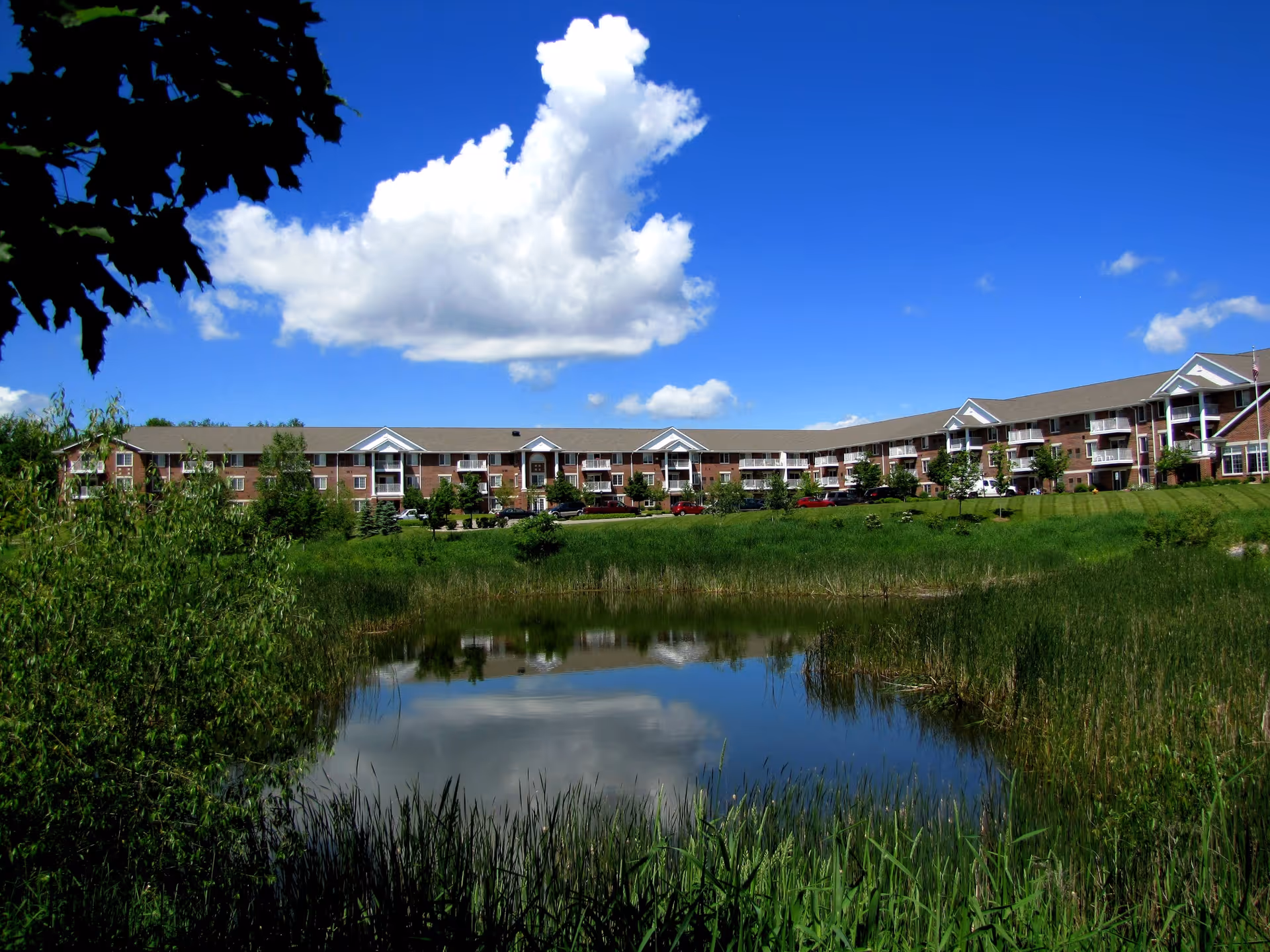 A long three-story brick apartment building reflected in a pond under a bright blue sky with a large white cloud.