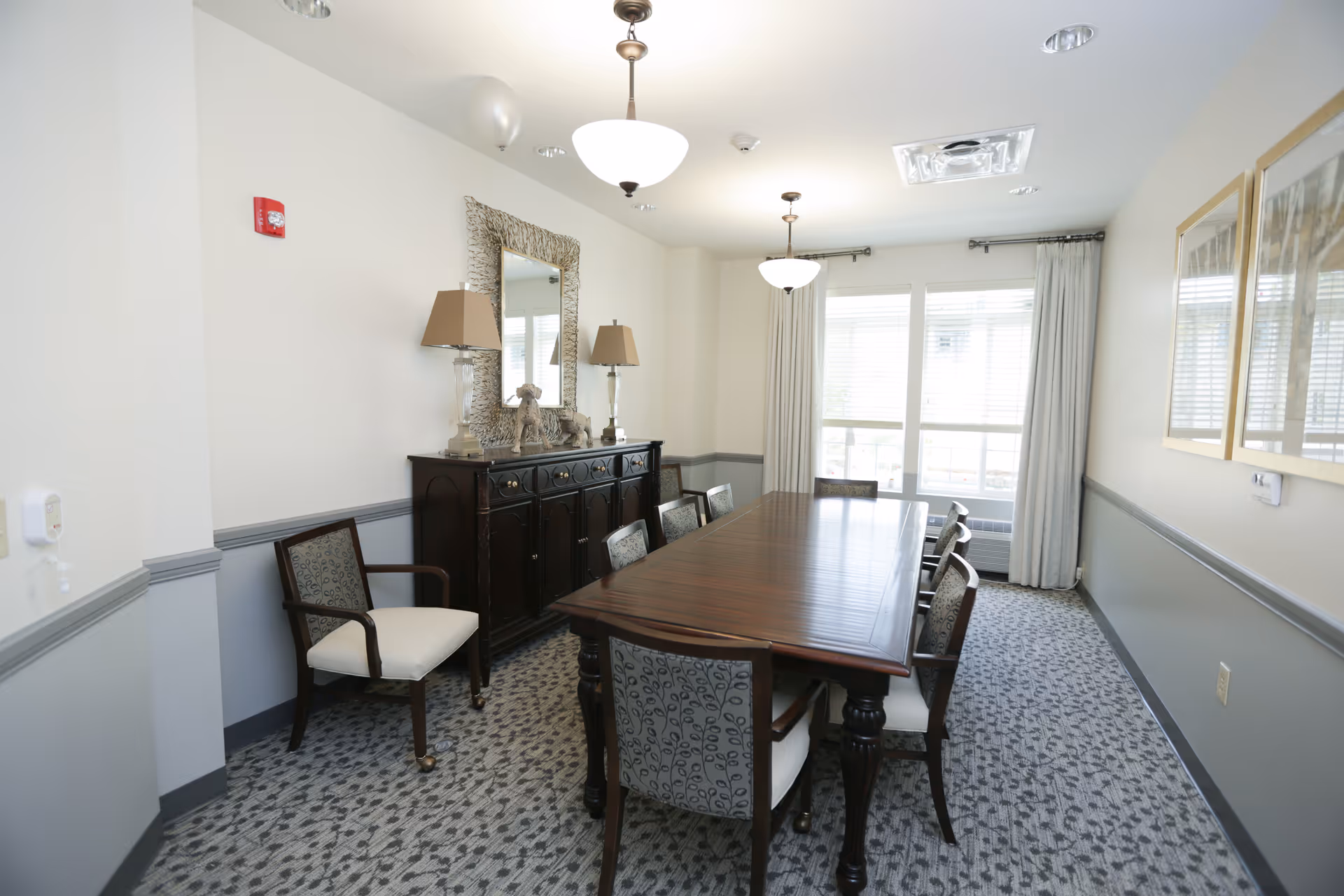A well-lit dining room with a long wooden table surrounded by eight upholstered chairs. The room features two hanging light fixtures, a large window with curtains, a dark wooden sideboard with two lamps and decorative items, a large mirror above the sideboard, and framed artwork on the wall. The walls are painted in a two-tone style with white upper walls and gray lower walls, and the floor is carpeted with a patterned design.