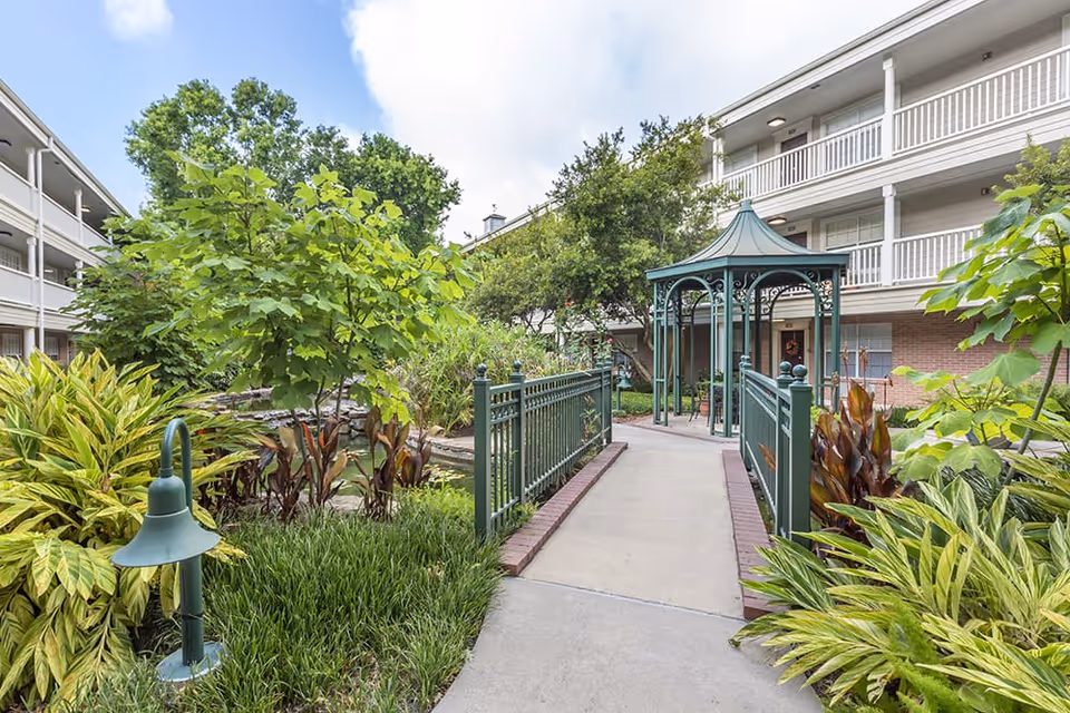 Outdoor garden area at Brookdale West University featuring a paved walkway with green metal railings leading to a decorative gazebo. The garden is lush with various green plants and trees, surrounded by a multi-story building with balconies.